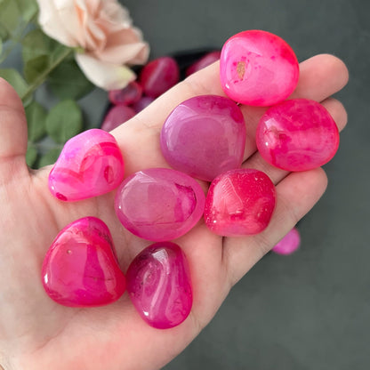 A wooden bowl filled with smooth, polished Pink Onyx Tumbled Stones of varying sizes and shades. The stones have a glossy finish and some exhibit white marbling, offering a mix of vibrant and subtle pink hues. The background is white, making the colors pop.