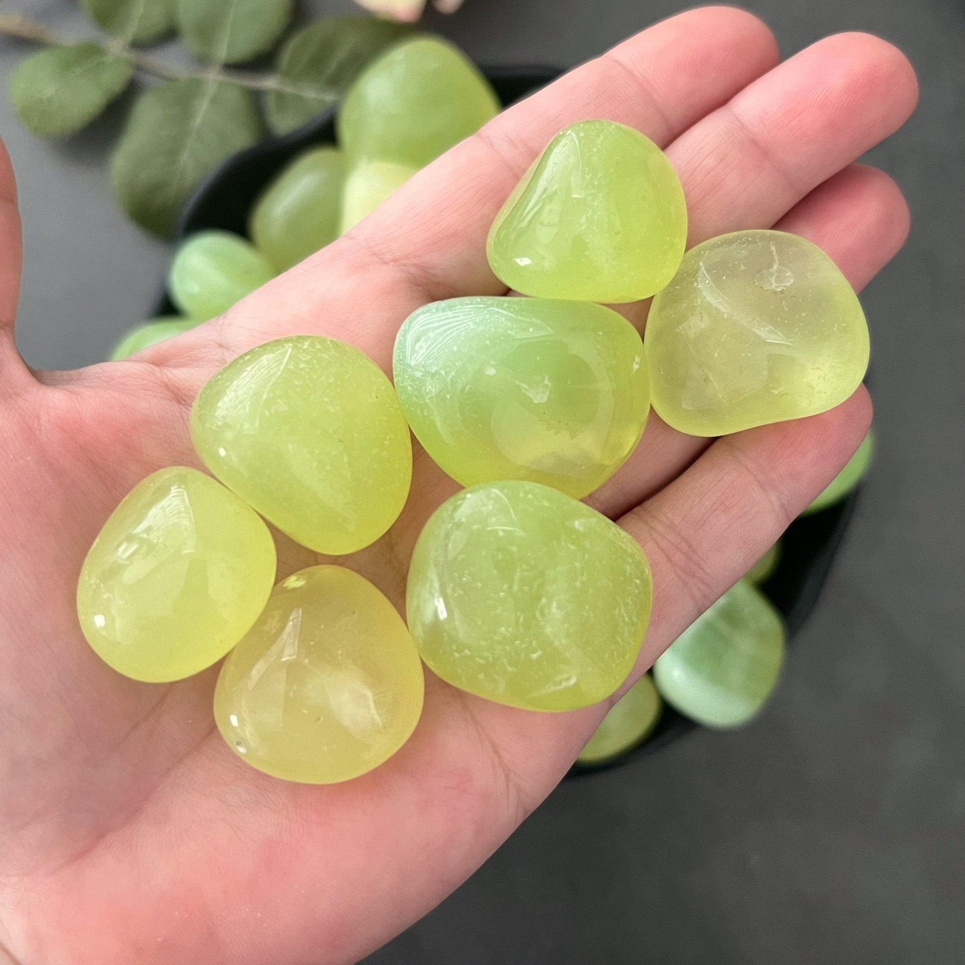 A wooden bowl filled with an assortment of polished, light green stones, including Parrot Green Onyx Tumbled Stones for anxiety relief, arranged on a white background. The stones have a smooth, shiny surface and varying sizes.