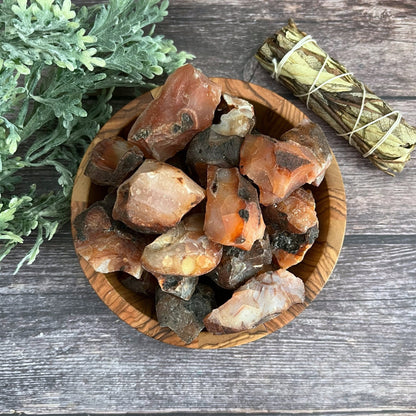 A wooden bowl full of rough, unpolished crystal stones sits on a wooden surface. To the left of the bowl is a sprig of leafy green plant, and above the bowl is a bundle of sage wrapped in twine. Among the crystals, vibrant Raw Carnelian Stones stand out, radiating motivation and endurance.