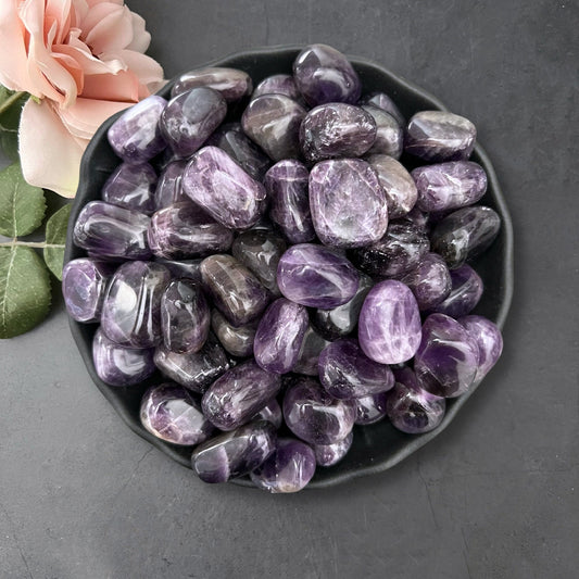 A black plate filled with polished Dark Dream Amethyst Tumbled Stones is placed on a dark surface. To the left of the plate, there are light pink rose flowers with green leaves. The amethyst stones, intuitively chosen for their stress-relieving properties, have a glossy finish and exhibit various shades of purple.