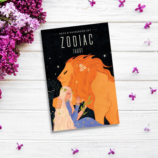 A "Zodiac Tarot" sits on a white wooden surface surrounded by purple and pink flowers, ideal for those passionate about astrology. The cover features an illustration of a lion and a woman with long blonde hair holding a flower.