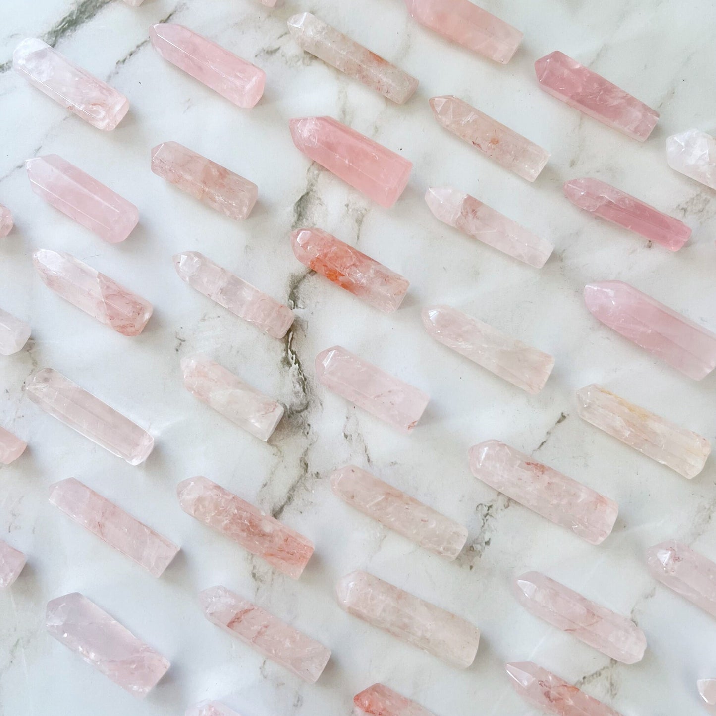 A wooden bowl filled with 1-2" Rose Quartz Mini Point is placed on a white marble surface. A pink rose with green leaves lies beside the bowl, adding a touch of elegance to the serene scene, symbolizing unconditional love.