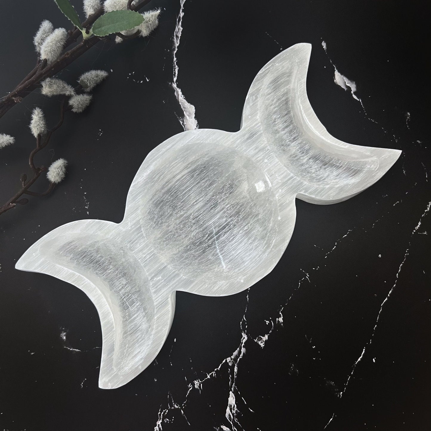 A white, candy-shaped selenite crystal sits on a dark surface, cradled in a polished 8" Selenite Triple Moon Bowl and surrounded by various stones of different colors and textures. Pussy willow branches add a touch of nature to the arrangement, perfect for enhancing spiritual practices.