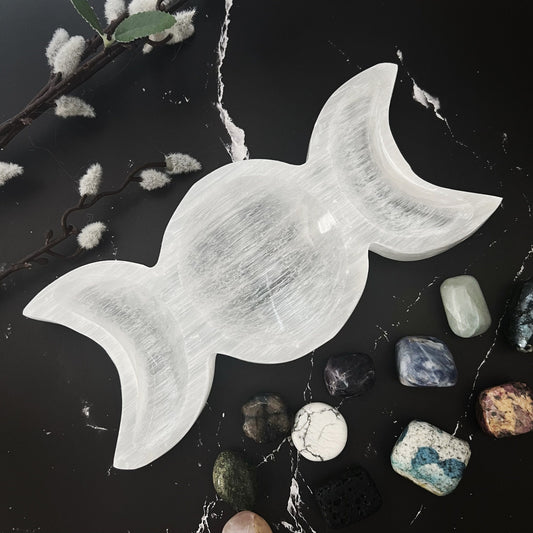 A white, candy-shaped selenite crystal sits on a dark surface, cradled in a polished 8" Selenite Triple Moon Bowl and surrounded by various stones of different colors and textures. Pussy willow branches add a touch of nature to the arrangement, perfect for enhancing spiritual practices.