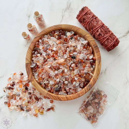 A wooden bowl filled with various tumbled and polished stones sits on a marble surface. Surrounding the bowl are three small glass bottles containing Hematoid Quartz, a bundle of red sage, and a plastic bag with Fire Quartz Crystal Chips.