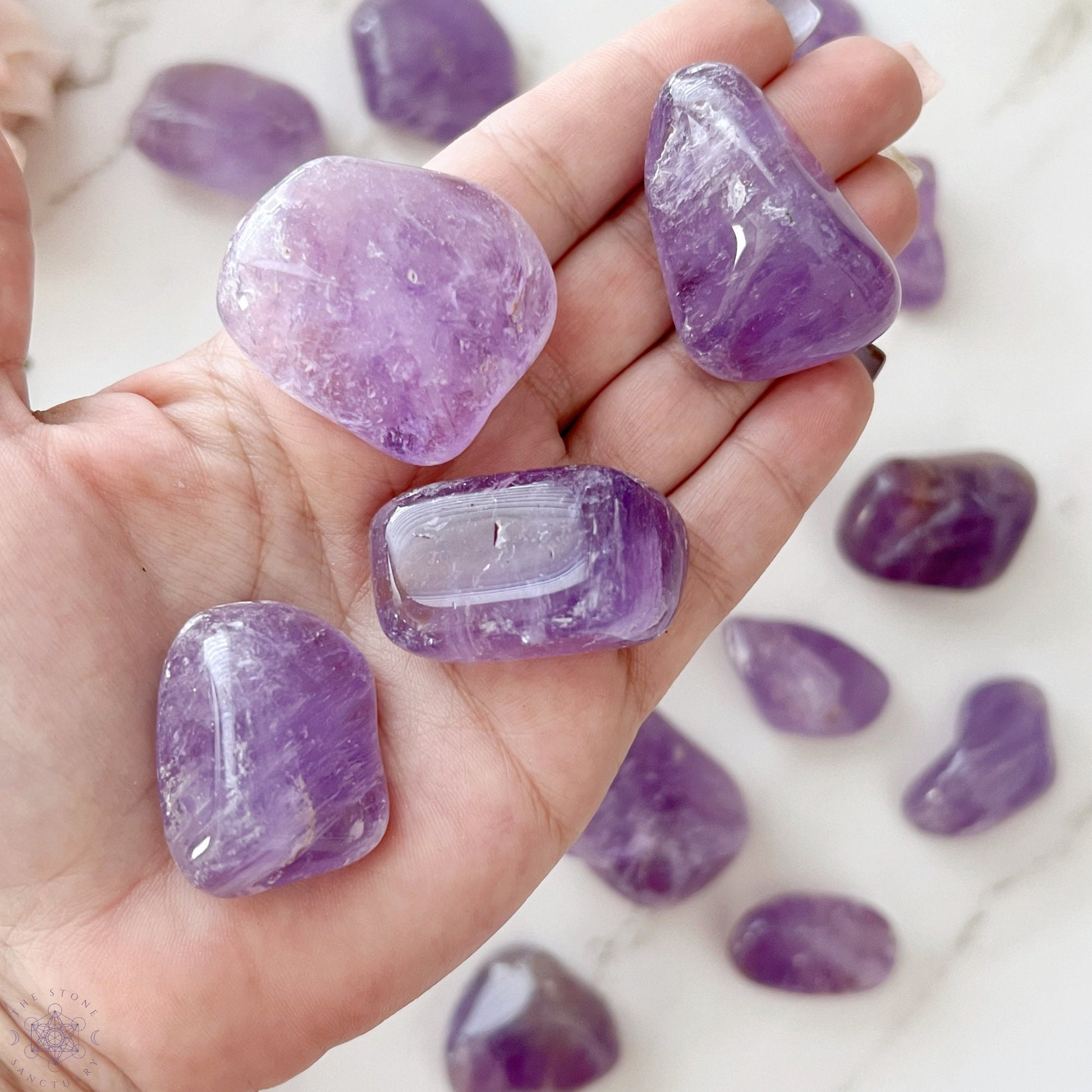 A wooden bowl filled with Amethyst Tumbled Stones (Brazil) is displayed on a white background. These smooth, shiny stones, in varying shades of purple, are known for their psychic protection and ability to relieve stress. The bowl itself has a natural wood grain pattern that complements the beauty of the amethysts.