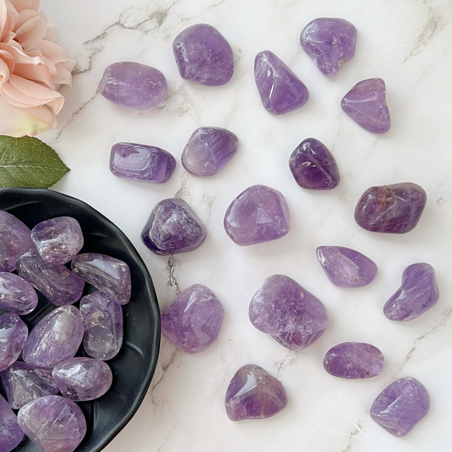 A wooden bowl filled with Amethyst Tumbled Stones (Brazil) is displayed on a white background. These smooth, shiny stones, in varying shades of purple, are known for their psychic protection and ability to relieve stress. The bowl itself has a natural wood grain pattern that complements the beauty of the amethysts.