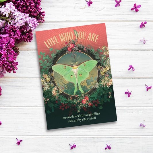 A beautifully illustrated book placed on a white wooden surface surrounded by scattered small purple flowers and lilac blossoms. The book's cover reads "Love Who You Are Oracle," featuring a central image of a green moth amidst lush florals, inspiring confidence and self-worth.