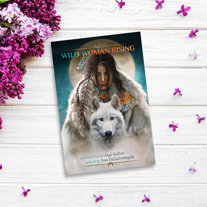 A book titled *Wild Woman Rising* by Angi Sullins, accompanied by the *Wild Woman Rising Oracle*, is placed on a white wooden surface. The cover features a woman with tribal markings and a wolf. Purple and white flowers surround the 104-page guidebook and its 44 cards on the table.