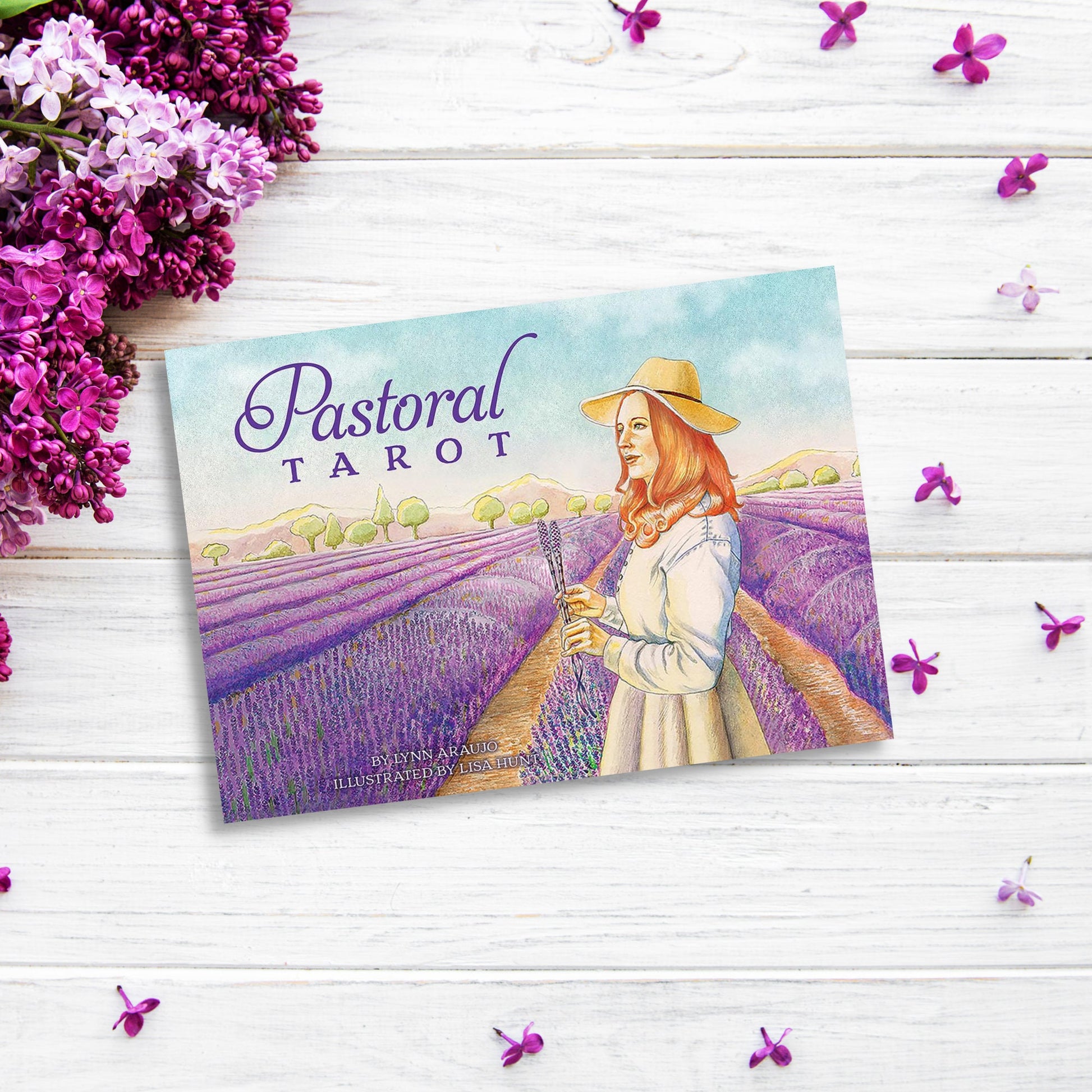 An image of a deck of "Pastoral Tarot" cards placed on a white wooden surface. The cover shows an illustration of a woman in a hat standing in a lavender field, with lilac flowers and small purple petals surrounding the deck.