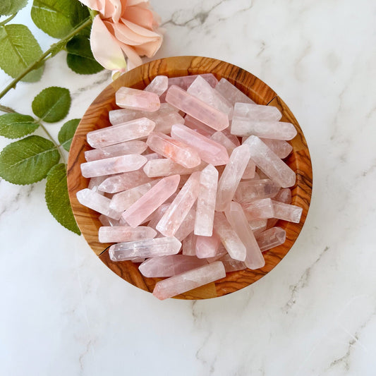 A wooden bowl filled with 1-2" Rose Quartz Mini Point is placed on a white marble surface. A pink rose with green leaves lies beside the bowl, adding a touch of elegance to the serene scene, symbolizing unconditional love.