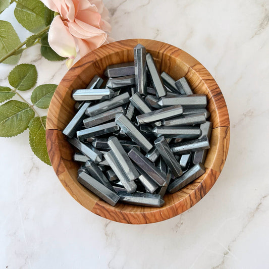 A wooden bowl filled with 1-2" Mini Hematite Point sits on a light marble surface, emanating grounding energy. To the left of the bowl, there is a pink rose with green leaves.