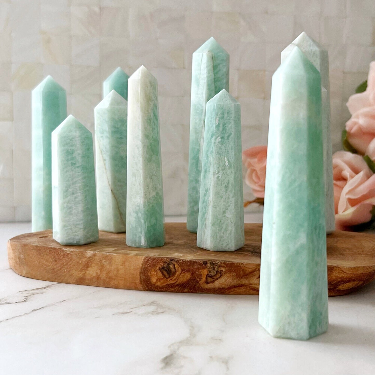 A group of light green crystal towers arranged on a wooden slab, highlighted by a 3-4" Amazonite Standing Point standing tall. The crystals of varying heights and pointed tops are complemented by blurred pink flowers in the background against a white, textured backdrop. The setting rests elegantly on a marble surface.