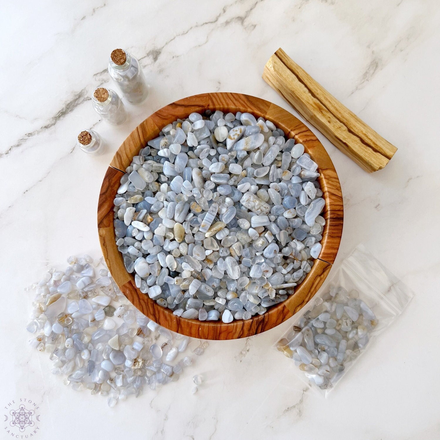 A wooden bowl filled with Blue Lace Agate Crystal Chips and white tumbled stones sits on a marble surface. Around the bowl are small glass bottles with corks containing more crystal chips, a small wooden stick, a spilled pile of stones, and a plastic bag with even more stones.