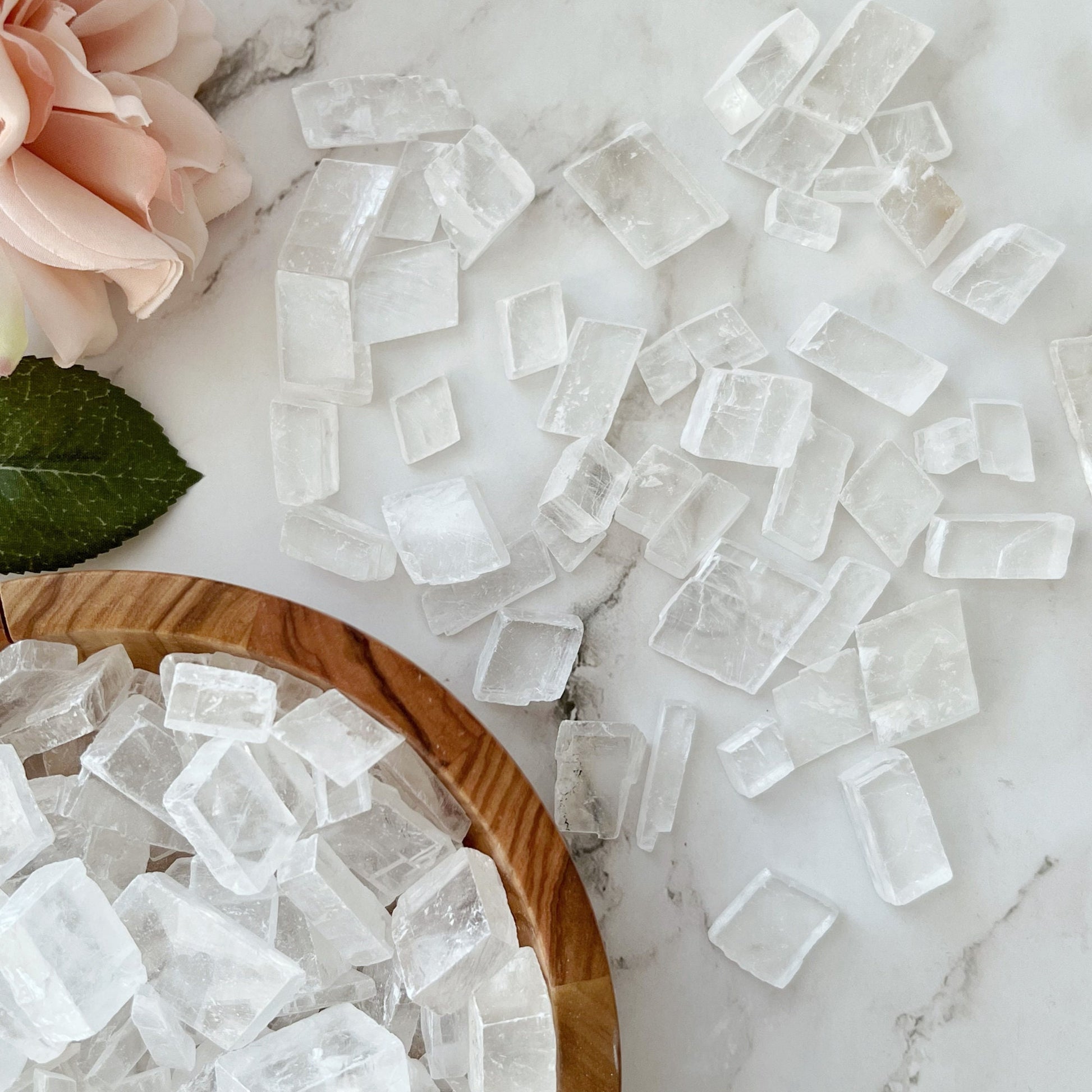 A wooden bowl filled with large, Optical Calcite Crystal Chips sits on a white marble surface. Beside the bowl, there is a pink rose with green leaves. The composition gives a natural and elegant feel, hinting at metaphysical uses of these raw stones.