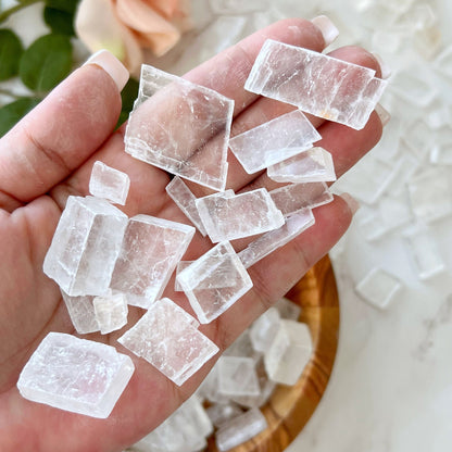 A wooden bowl filled with large, Optical Calcite Crystal Chips sits on a white marble surface. Beside the bowl, there is a pink rose with green leaves. The composition gives a natural and elegant feel, hinting at metaphysical uses of these raw stones.