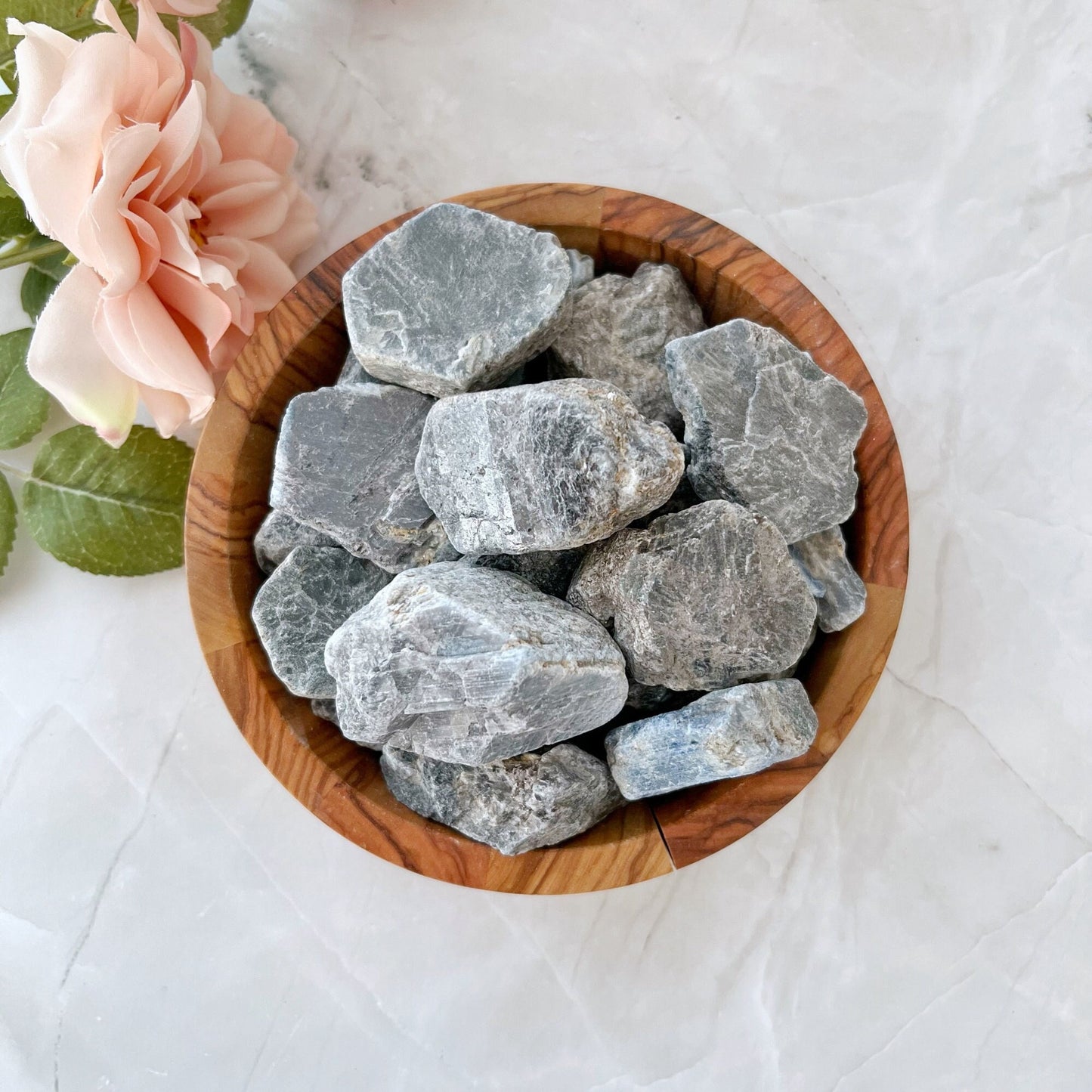 A wooden bowl filled with Raw Sapphire Stones sits on a white marble surface. A pink rose with green leaves is positioned to the left of the bowl, adding a touch of natural elegance to the scene, while one protective stone suggests a sense of serenity and spiritual truth to the arrangement.