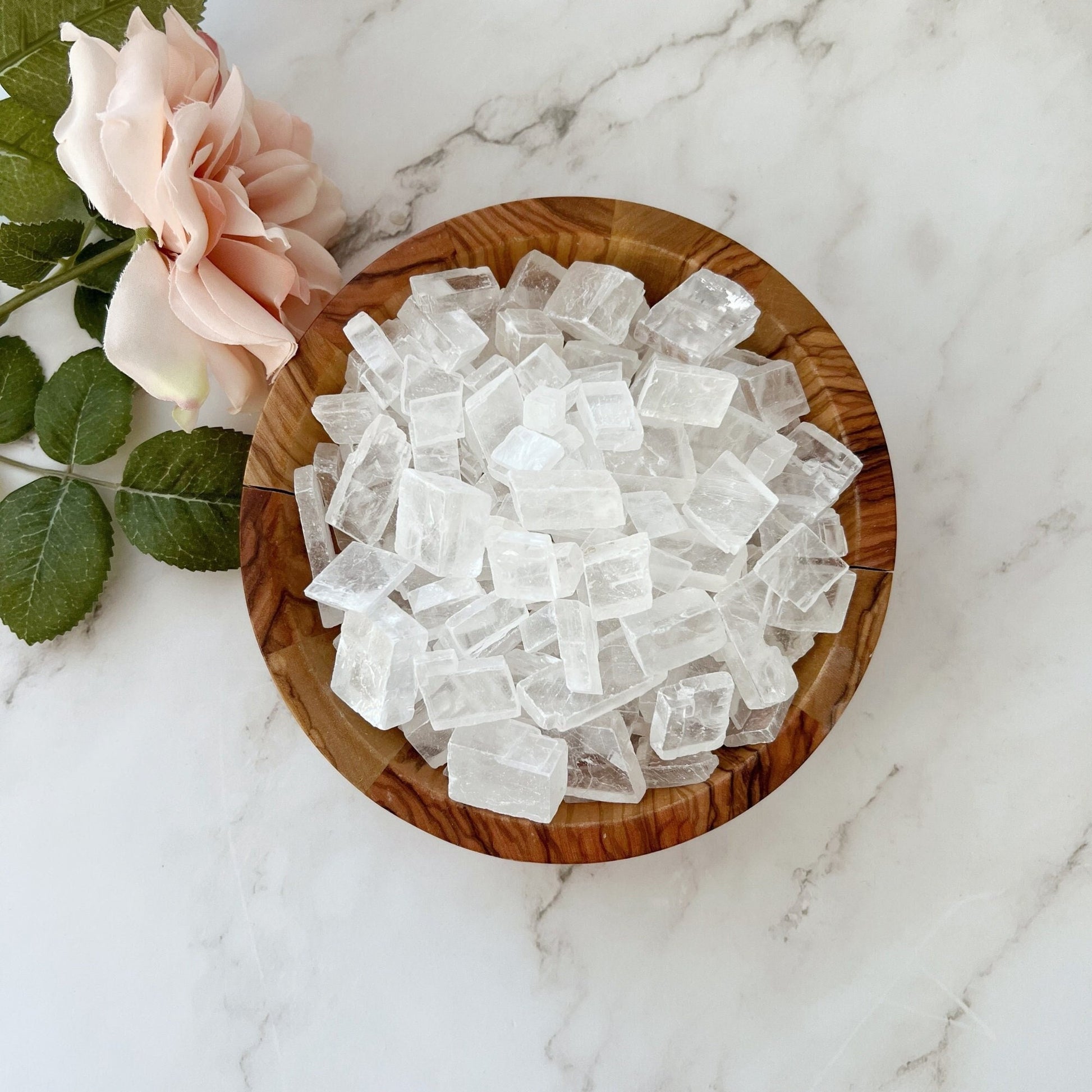 A wooden bowl filled with large, Optical Calcite Crystal Chips sits on a white marble surface. Beside the bowl, there is a pink rose with green leaves. The composition gives a natural and elegant feel, hinting at metaphysical uses of these raw stones.