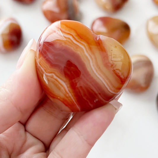 Close-up of a hand holding a polished, heart-shaped Banded Carnelian Agate Heart Palm Stone with orange, white, and brown bands. Several similar polished stones are blurred in the background. The stone's surface is glossy, reflecting light and showing intricate patterns.