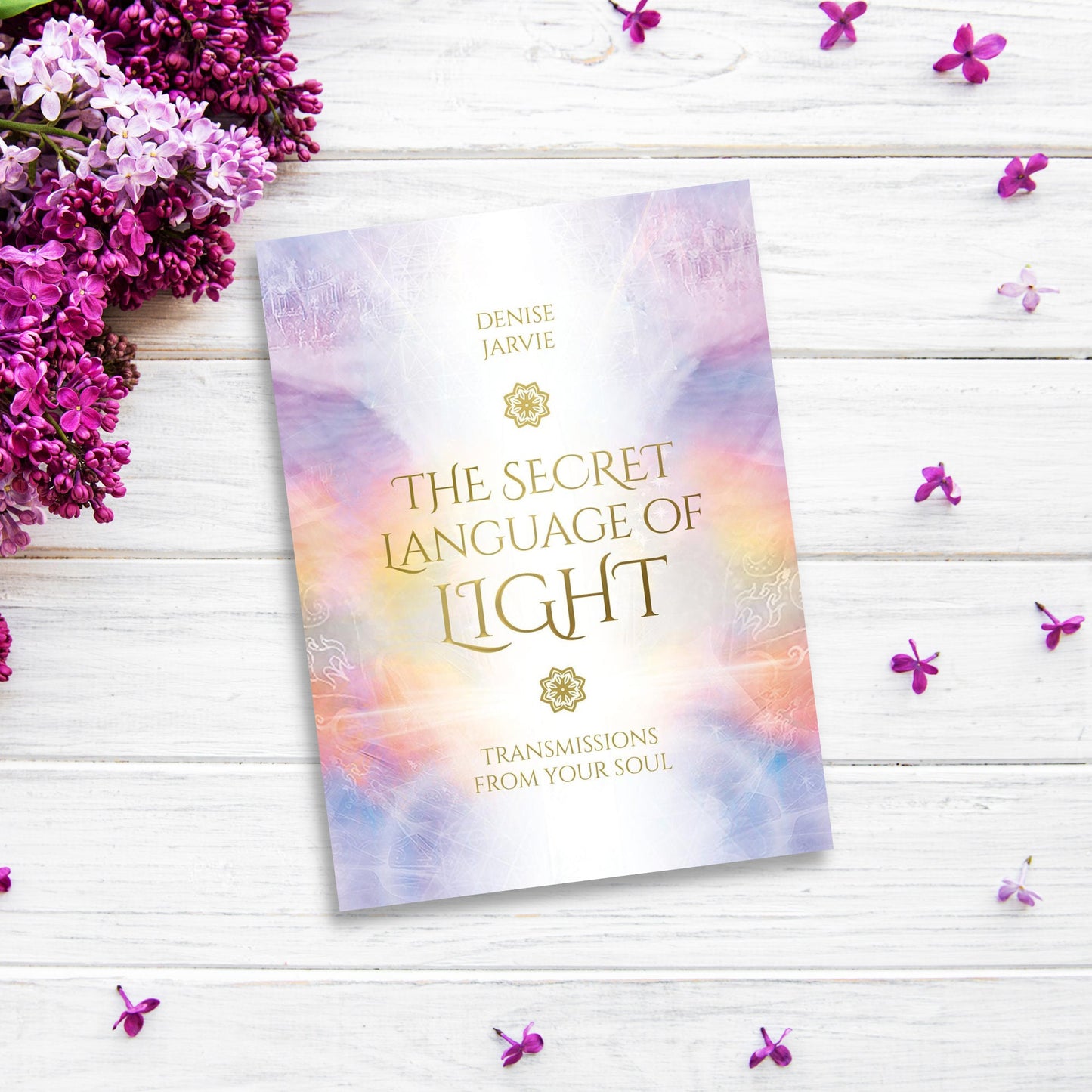 A book titled "The Secret Language of Light Oracle" by Denise Jarvie is placed on a white wooden surface. An Oracle deck lies beside it, surrounded by clusters of vibrant purple flowers and scattered petals that add a decorative touch.