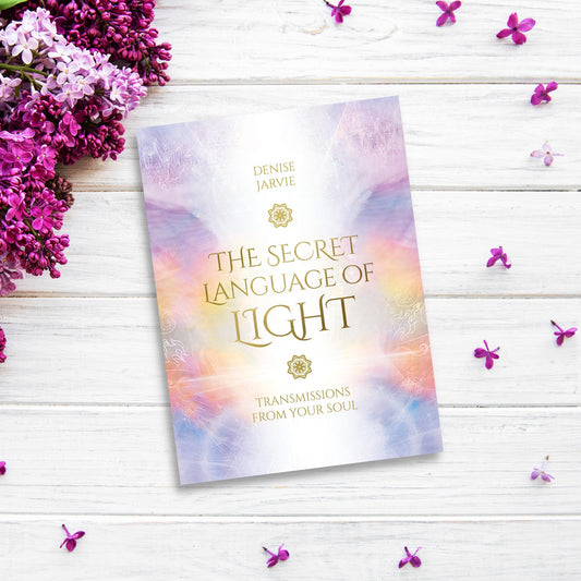 A book titled "The Secret Language of Light Oracle" by Denise Jarvie is placed on a white wooden surface. An Oracle deck lies beside it, surrounded by clusters of vibrant purple flowers and scattered petals that add a decorative touch.