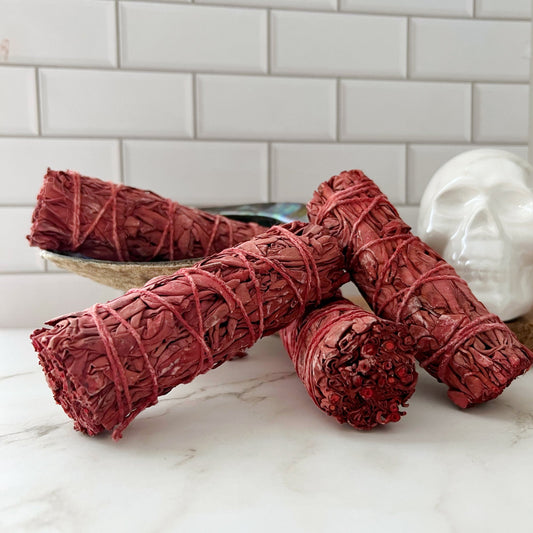 Four Organic Dragon's Blood White Sage Bundles tied with string rest on a white marble surface. They are arranged in front of a white ceramic skull decor piece and a shallow bowl, with a white tiled wall in the background.