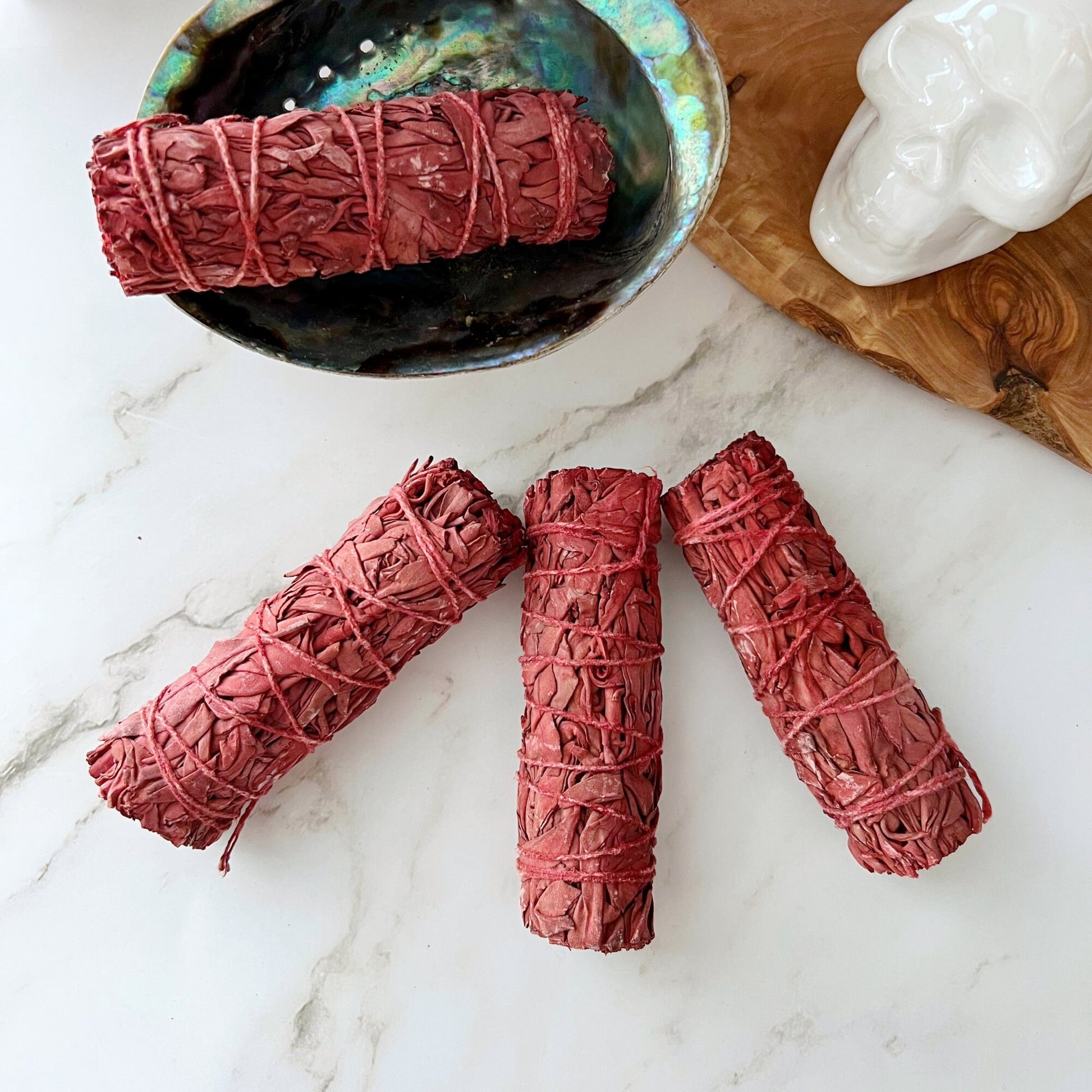 Four Organic Dragon's Blood White Sage Bundles tied with string rest on a white marble surface. They are arranged in front of a white ceramic skull decor piece and a shallow bowl, with a white tiled wall in the background.