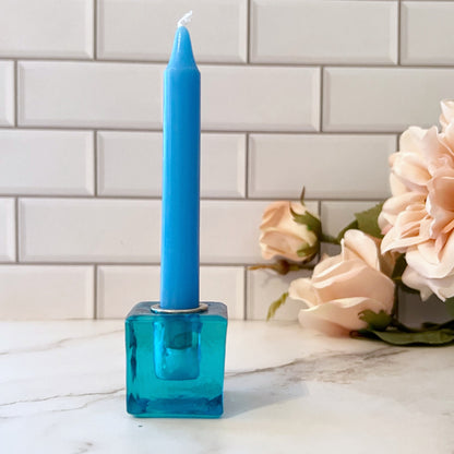 A set of eight colorful candles, each in a 1" Mini Glass Cube Holder for Ritual Candles of matching color, arranged in a line on a white tiled surface. Colors from left to right are: deep purple, dark blue, teal, light blue, green, yellow, orange, and red.