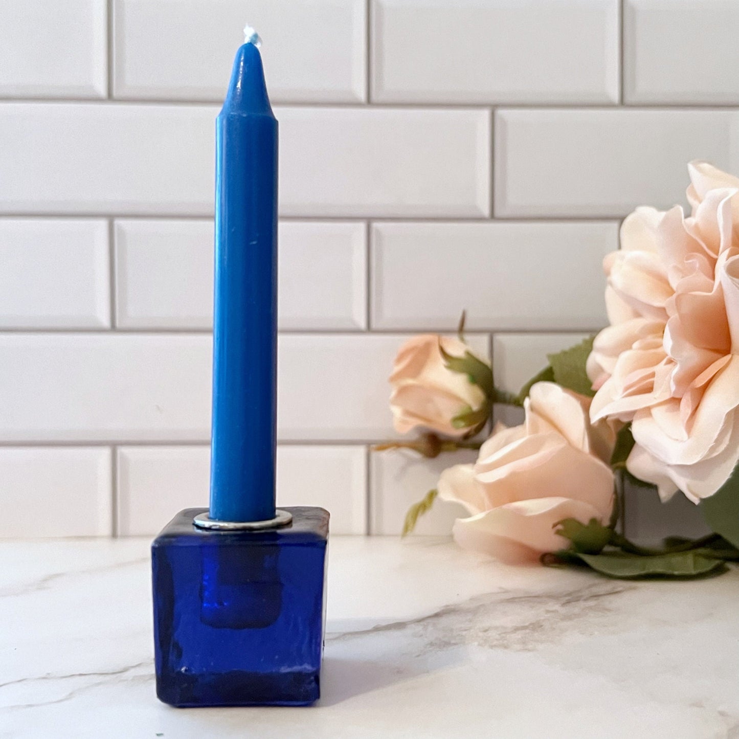 A set of eight colorful candles, each in a 1" Mini Glass Cube Holder for Ritual Candles of matching color, arranged in a line on a white tiled surface. Colors from left to right are: deep purple, dark blue, teal, light blue, green, yellow, orange, and red.