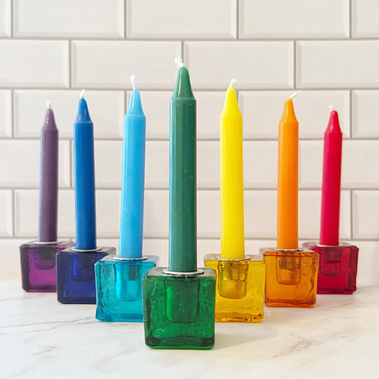 A set of eight colorful candles, each in a 1" Mini Glass Cube Holder for Ritual Candles of matching color, arranged in a line on a white tiled surface. Colors from left to right are: deep purple, dark blue, teal, light blue, green, yellow, orange, and red.