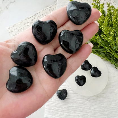 A collection of small, polished 1" Mini Black Tourmaline Pocket Heart stones are arranged on a light circular surface, with more of the same stones in a golden dish next to it. Green foliage and textured paper are visible in the background, offering negative energy protection.