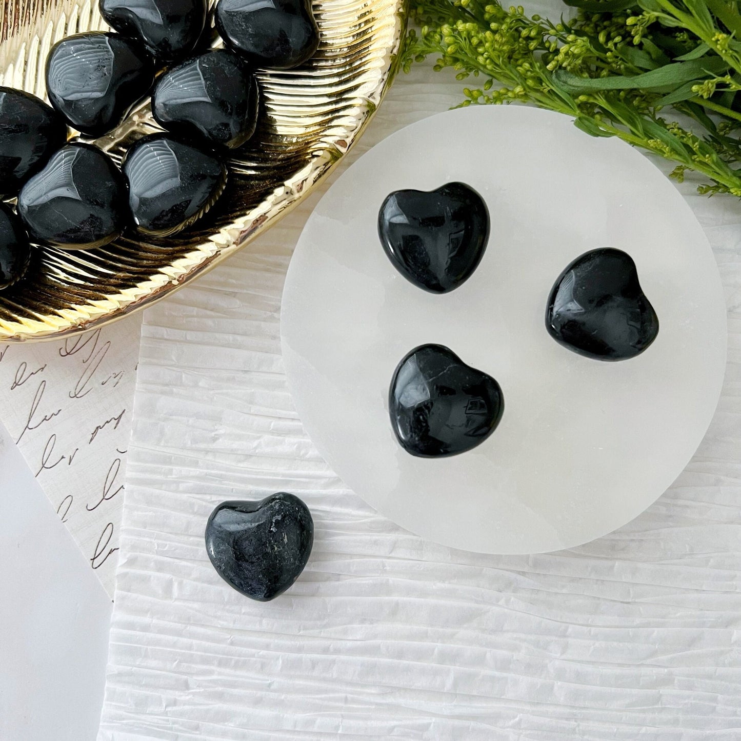 A collection of small, polished 1" Mini Black Tourmaline Pocket Heart stones are arranged on a light circular surface, with more of the same stones in a golden dish next to it. Green foliage and textured paper are visible in the background, offering negative energy protection.