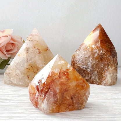 Three polished, pyramid-shaped crystals with hues of white, orange, and brown are displayed on a textured white surface. In the background, a soft pink rose is visible on the left side against a light gray backdrop. Among them, the 2-4" Golden Healer Quartz Top Polished Point shines brightly, enhancing spiritual healing and connecting to the Crown Chakra.