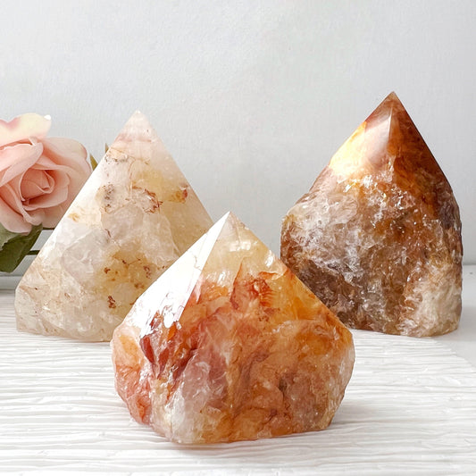 Three polished, pyramid-shaped crystals with hues of white, orange, and brown are displayed on a textured white surface. In the background, a soft pink rose is visible on the left side against a light gray backdrop. Among them, the 2-4" Golden Healer Quartz Top Polished Point shines brightly, enhancing spiritual healing and connecting to the Crown Chakra.