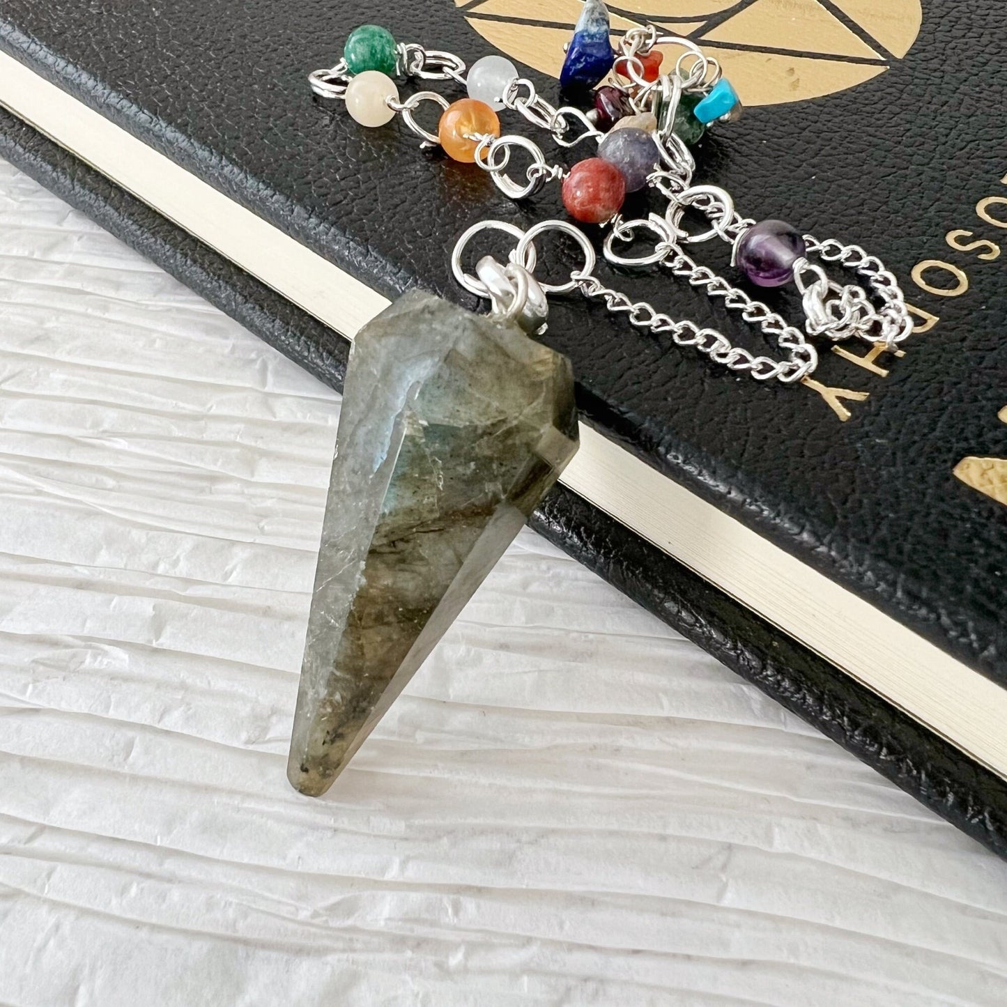 A Labradorite Hexagonal Pendulum attached to a silver chain featuring various colorful chakra crystals rests atop a black book with a gold geometric pattern on a white textured surface, serving as an exquisite divination tool.