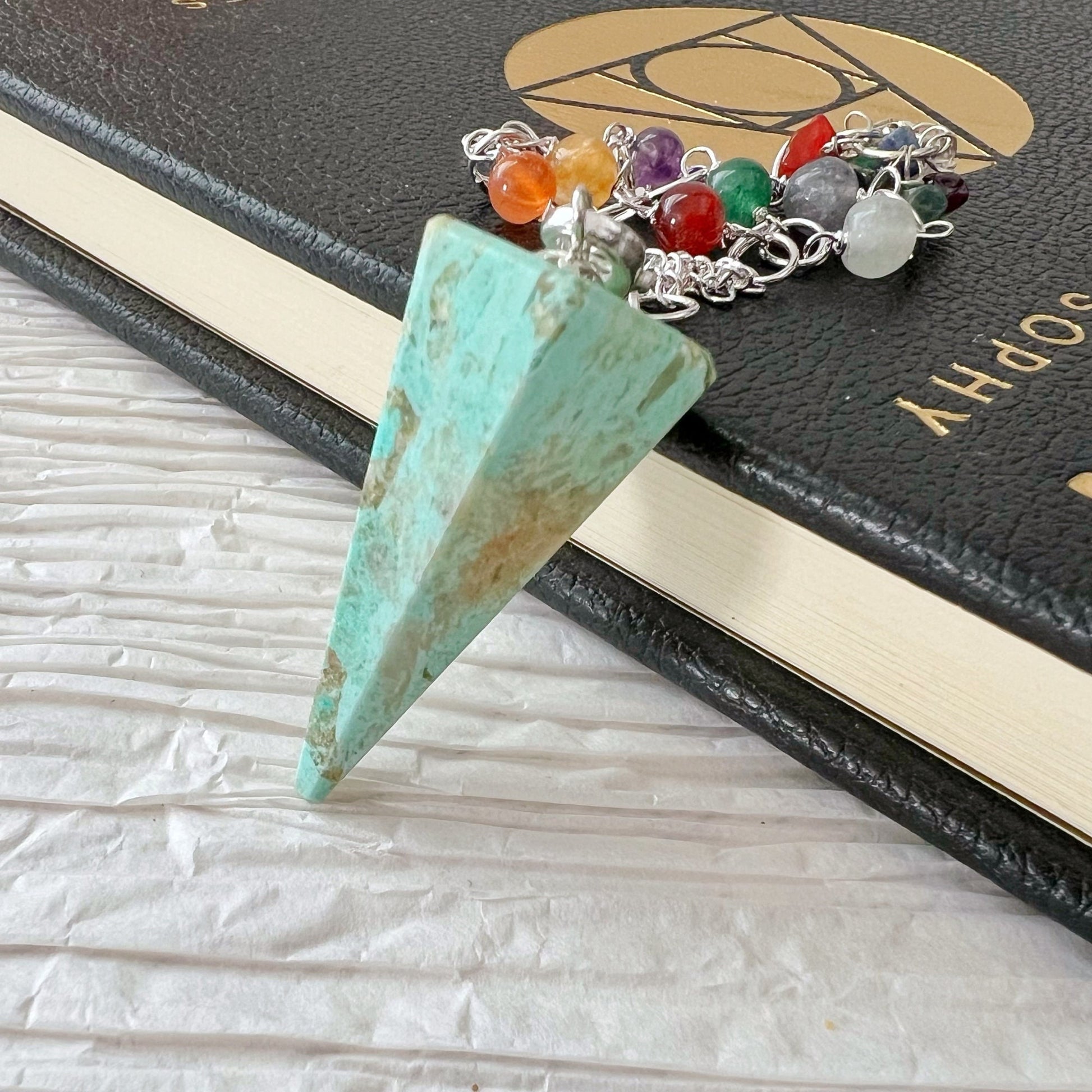 A Peruvian Turquoise Hexagonal Pendulum rests on the edge of a black book. The pendulum, often used in divination work, is connected to a Chakra crystal chain adorned with small, colorful stones. The background is a textured, light-colored surface.