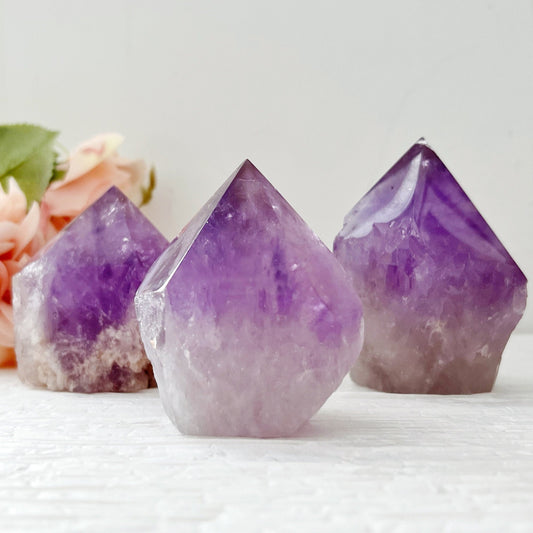 Three 2-4" Amethyst Top Polished Points with a gradient of clear to deep purple stand on a white surface. These natural Amethyst stones have pointed tops and rough bases. In the background, soft pink flowers are slightly out of focus, adding a touch of tranquility to this scene.