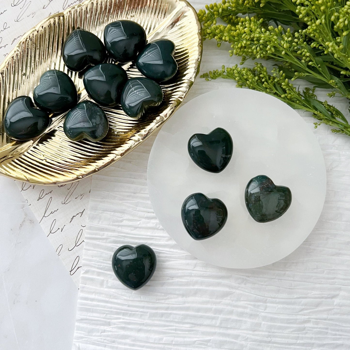 A set of small, polished 1" Mini Bloodstone Hearts displayed on a golden dish and a white circular platform. The background includes green foliage and a piece of paper with cursive text. These stones offer grounding and emotional support, with one heart-shaped stone placed beside the platform.