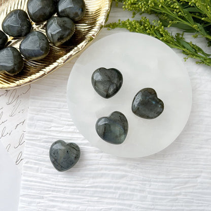 Displayed on a round white dish is a collection of 1" Mini Labradorite Crystal Hearts, each polished and heart-shaped. A golden bowl containing more Mini Labradorite hearts is positioned to the side, accompanied by greenery. The entire setting rests on a textured white surface with subtle hints of handwritten text in the background, creating an enchanting personal talisman decor.