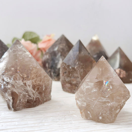 A close-up image features several 2-4" Smoky Quartz Top Polished Points, arranged meticulously in a crystal grid on a light-colored surface. The crystals vary in size and color intensity, with some exhibiting clear and reflective surfaces. Blurred flowers are visible in the background.