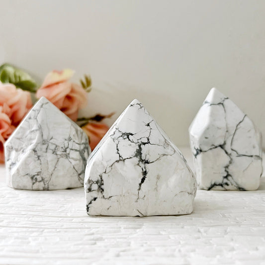 Three smooth, pointed 2-3" Howlite Top Polished Points are arranged on a textured white surface with soft peach-colored flowers in the blurred background, creating a serene and calming display.