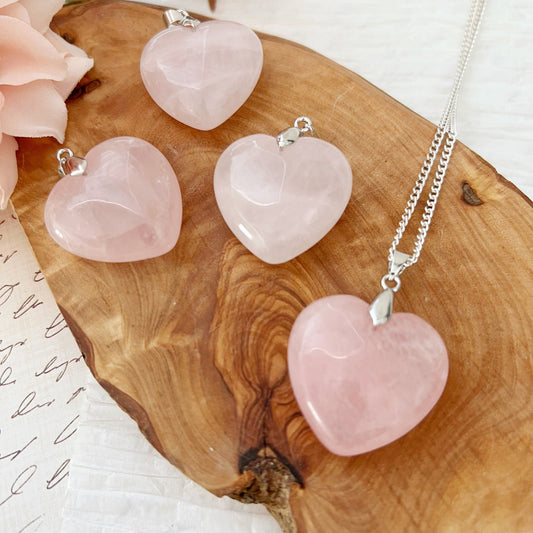 A photograph captures the essence of romantic love with four 1" Rose Quartz Crystal Heart Pendants resting on a wooden surface. Three lay flat, while one is attached to a silver necklace chain. A pink flower and a piece of paper with cursive writing are partly visible in the background, symbolizing emotional healing.