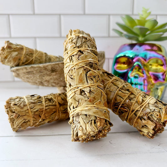 A close-up of Organic White Sage with Benzoin tied with string. They are resting on a white surface in front of a ceramic bowl. In the background, there is a colorful, iridescent skull decoration and a small potted succulent plant against a white tiled wall.