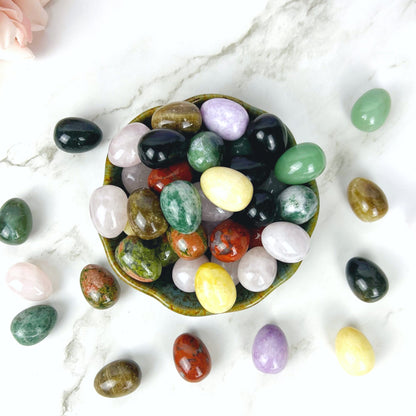 A collection of polished gemstone eggs, perfect for energy healing, is displayed on a light surface. Some 1" Mini Crystal Eggs are inside a greenish pottery bowl, while others are arranged around it. Pink roses are in the background, and the backdrop features soft bokeh lights.