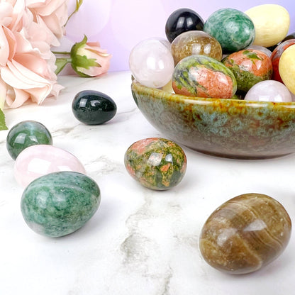 A collection of polished gemstone eggs, perfect for energy healing, is displayed on a light surface. Some 1" Mini Crystal Eggs are inside a greenish pottery bowl, while others are arranged around it. Pink roses are in the background, and the backdrop features soft bokeh lights.