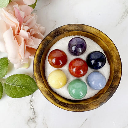 A wooden bowl filled with white sand holds a 7 Chakra Crystal Sphere Set. The spheres are arranged in a circle and include red, orange, yellow, green, blue, purple, and black stones. Near the bowl is a pink rose bloom and a green leaf on a white marble surface for energy healing.