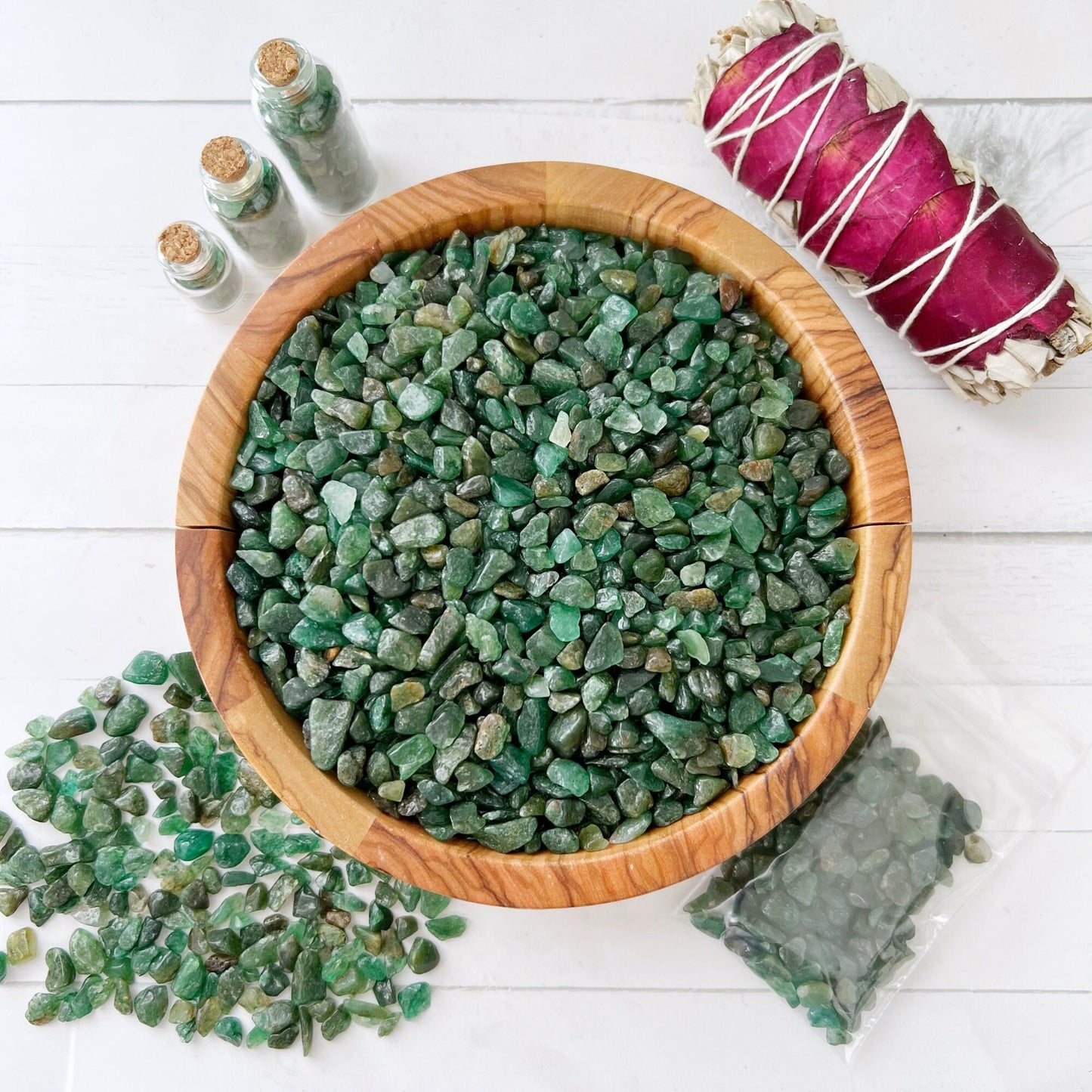 A wooden bowl filled with small, tumbled and polished green stones is centered on a white surface. Surrounding the bowl are mini bottles containing more green stones, a small bundle of sage wrapped in pink ribbon, and a plastic bag of additional Emerald Crystal Chips—perfect for creative and mystical endeavors.