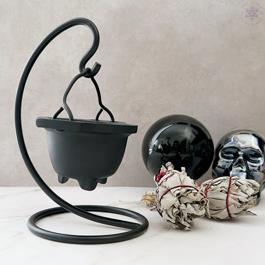 A Mini Cast Iron Cauldron with Stand, accompanied by two black orbs, one resembling a skull, and a bundle of sage wrapped with red string. This decorative piece is arranged on a light surface against a beige background, evoking the essence of magical practices.