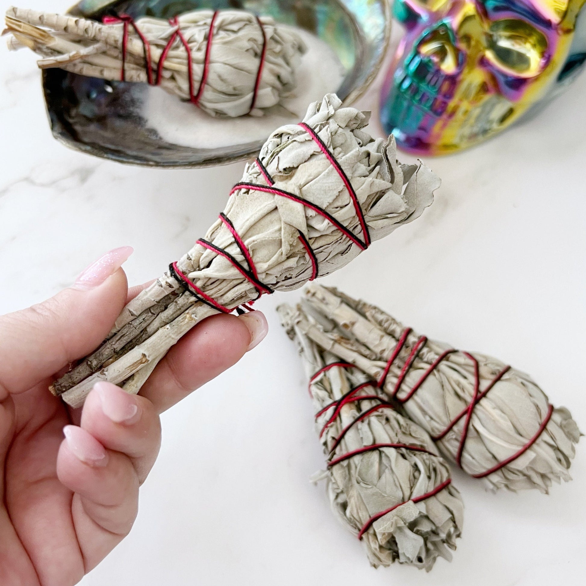 Bundles of Organic White Sage Torch Smudge Stick, tied with red string, are arranged on a white marble surface. A metallic rainbow-colored skull and a shiny shell dish add decorative elements to this cleansing and purifying scene.