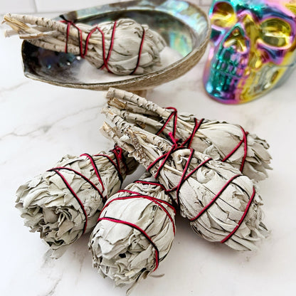 Bundles of Organic White Sage Torch Smudge Stick, tied with red string, are arranged on a white marble surface. A metallic rainbow-colored skull and a shiny shell dish add decorative elements to this cleansing and purifying scene.